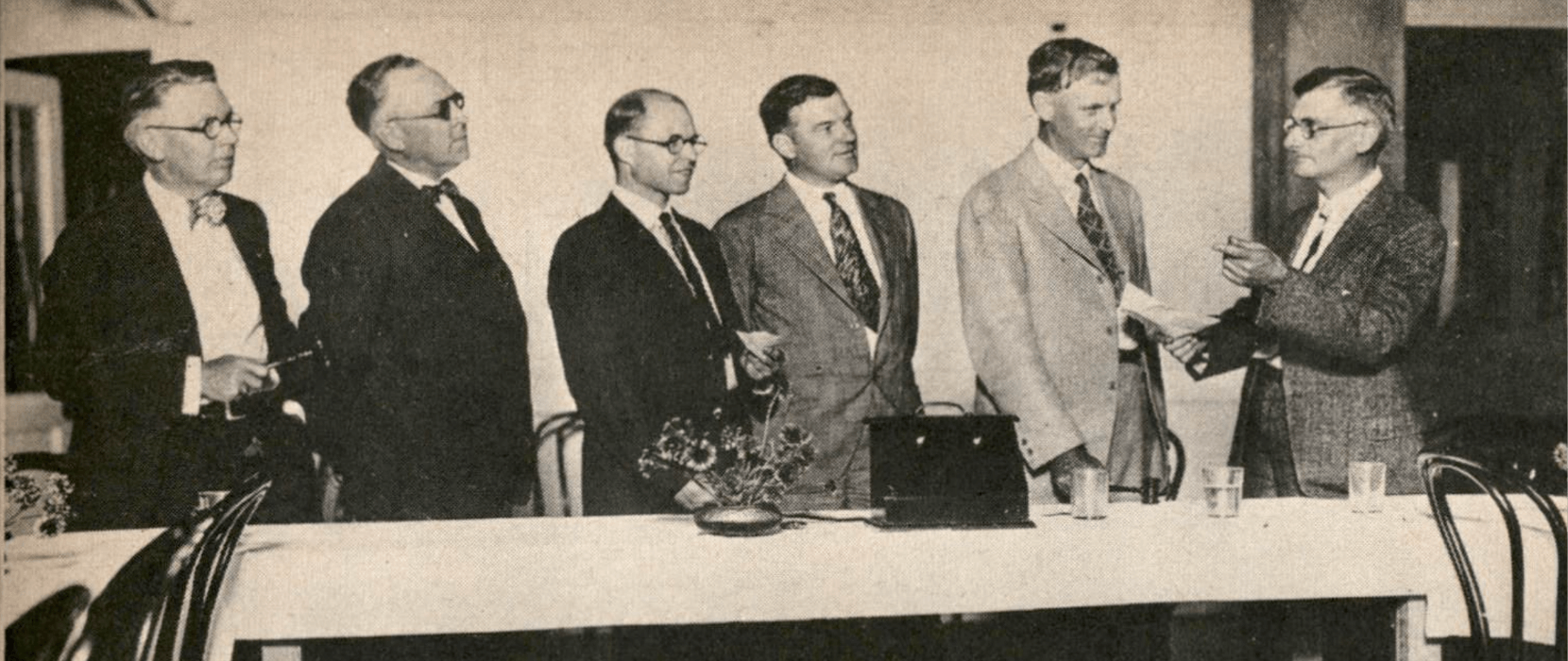 Black and white photo of six men standing behind a table