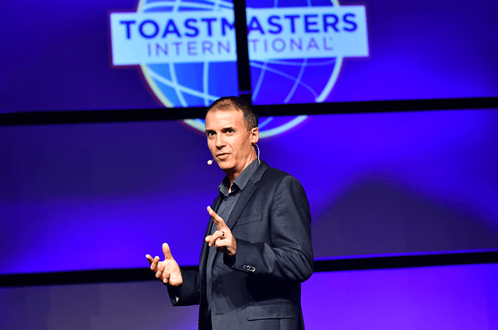 Man speaking onstage with blue background and Toastmasters International logo