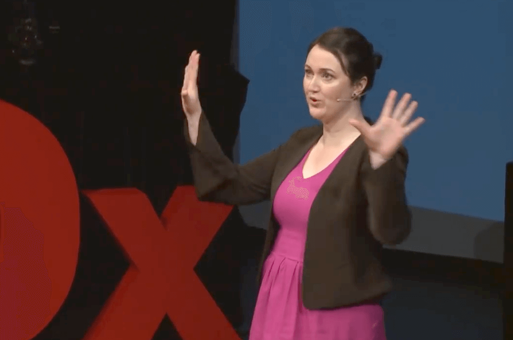 Woman in pink dress and black jacket using hand gestures while speaking on TEDX stage