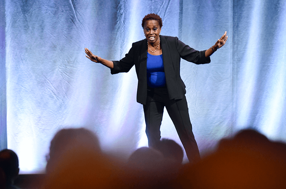 Woman in black suit and blue shirt speaking onstage using hand gestures