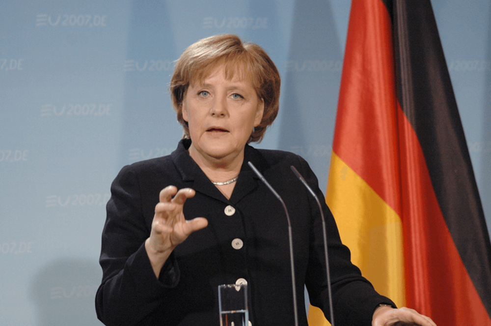 Angela Merkel in black outfit speaking into microphone near a German flag
