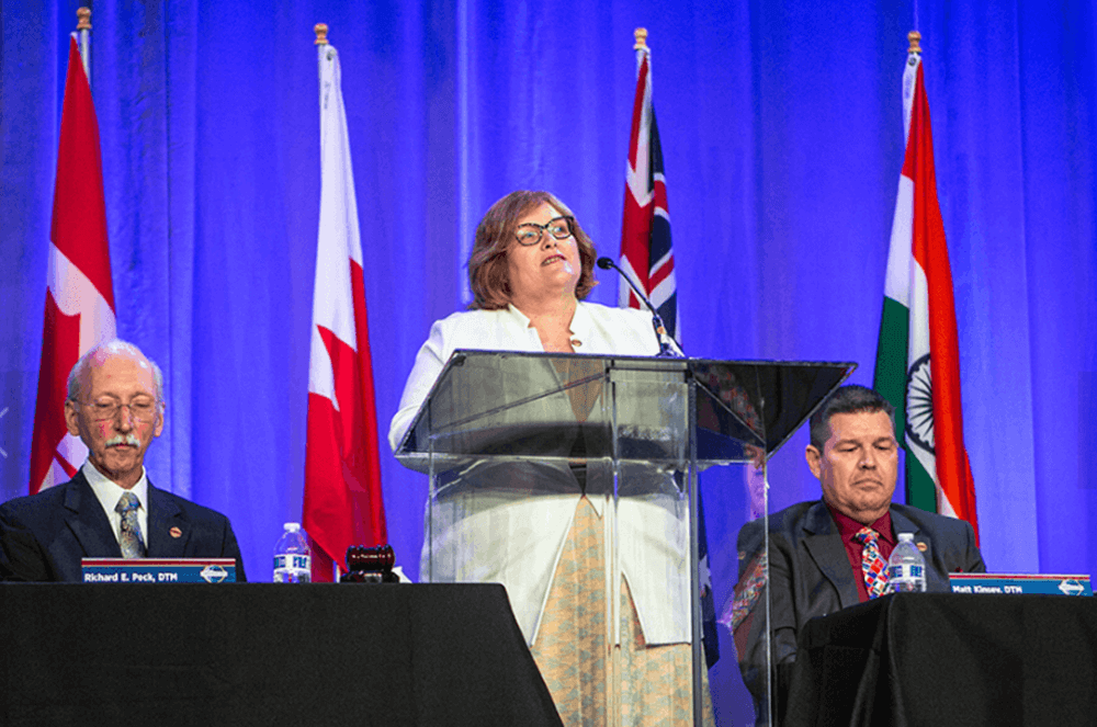Woman speaking at lectern onstage next to men at tables with flags in background