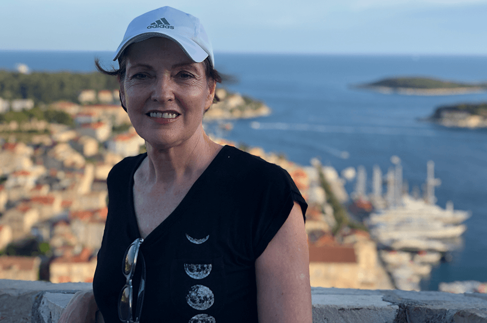Woman in short sleeved shirt and white hat posing with water and Croatian town in background